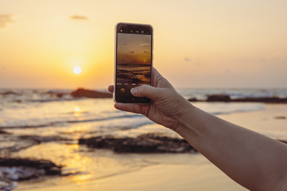 Woman using smart phone take a photo at evening on the beach of sunset