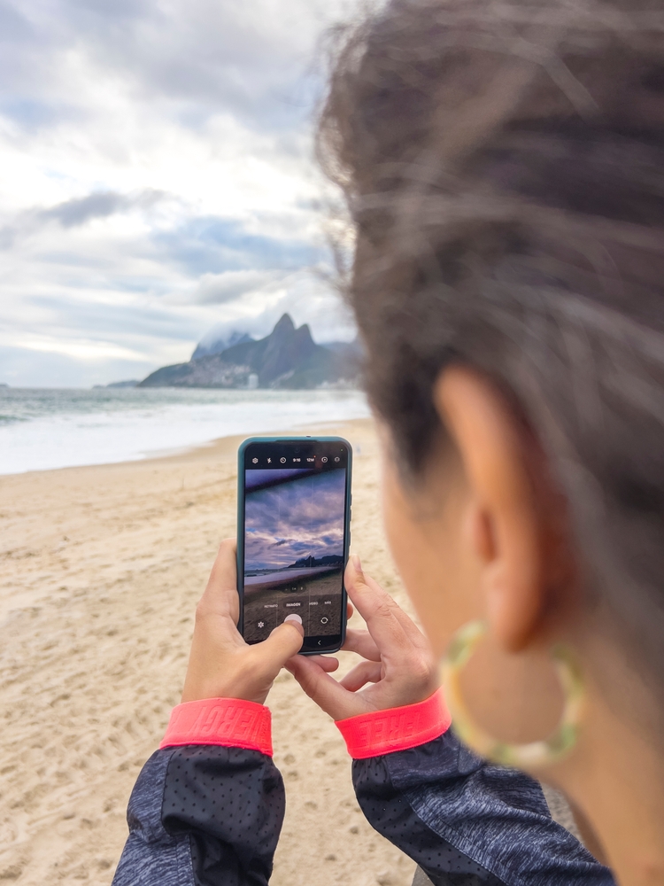 Woman photographing the waves and mountains of Ipanema Beach with her smartphone in Rio de Janeiro, Brazil. 