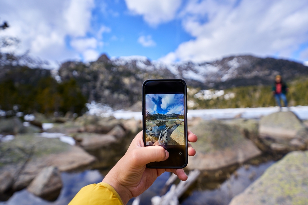 Hand holding phone taking a photo of a mountain landscape with snow