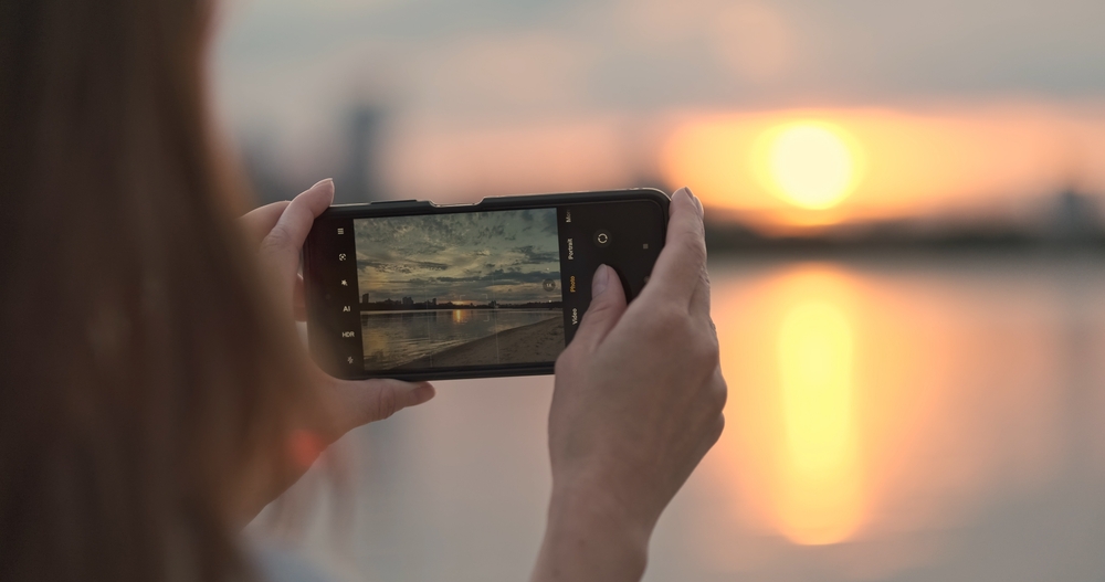 Girl takes smartphone photo of sunset on wide river.